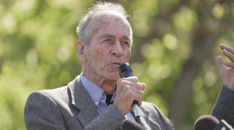 An older man with white hair and wearing a suit holds a microphone and stands outside.