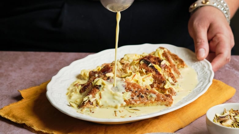 Closeup on a white platter with slices of Shahi Tukda, sweet, fried bread topped with rose petals and cardamom. A ladle at the top of the image is pouring a milky sauce over the dessert.