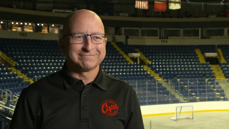 A man wearing a black shirt and glasses standing in an empty hockey arena. 