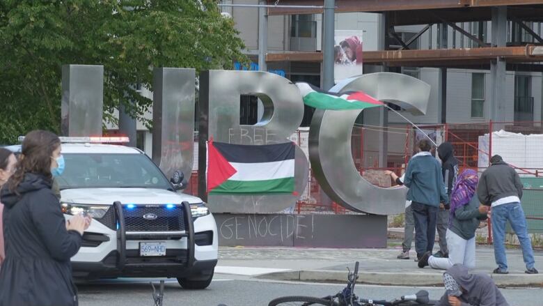 People fly Palestinian flags in front of a police car and a metal sign that says UBC.