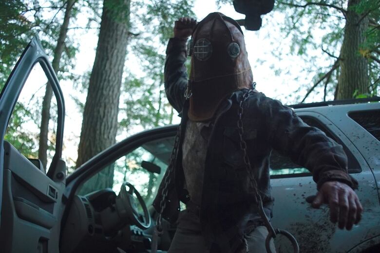 A man wearing a vintage firefighter's mask holds up an axe as if to strike someone. He is standing in front of a car spattered with blood. 