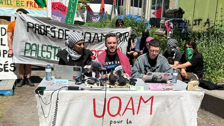 People speak into microphones while sitting at a table surrounded by banners.