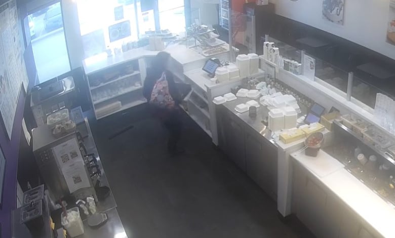 A still from a surveillance video shows a woman lifting a cash register from behind a counter.