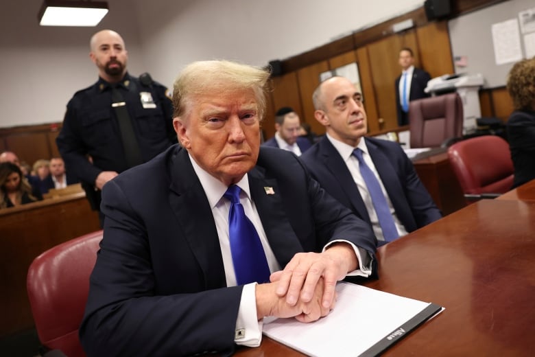 A man in a navy suit with a blue tie sits in a courtroom with his hands folded on the wooden desk in front of him.