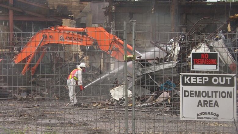 A worker in a hazardous materials suit is spraying debris with a hose. An excavator is working in the background. 