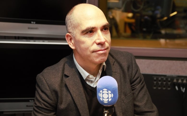 A balding man wearing a brown blazer speaks into a microphone.