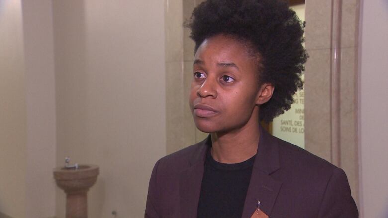 A non-binary person in a burgundy suit and black shirt stands in the hallway, outside the front door of an office.