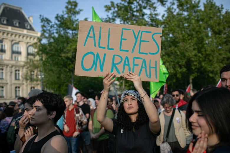 A woman wearing a Keffiyeh stands in a crowd using both hands to hold a cardboard sign over her head. The sign reads 'All eyes on Rafah' in blue lettering.