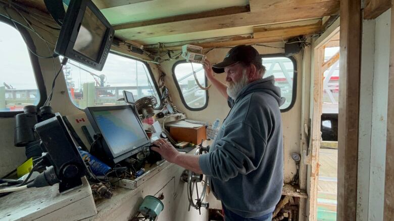 A white man with white beard, wearing a hat, working inside a boat's cockpit.