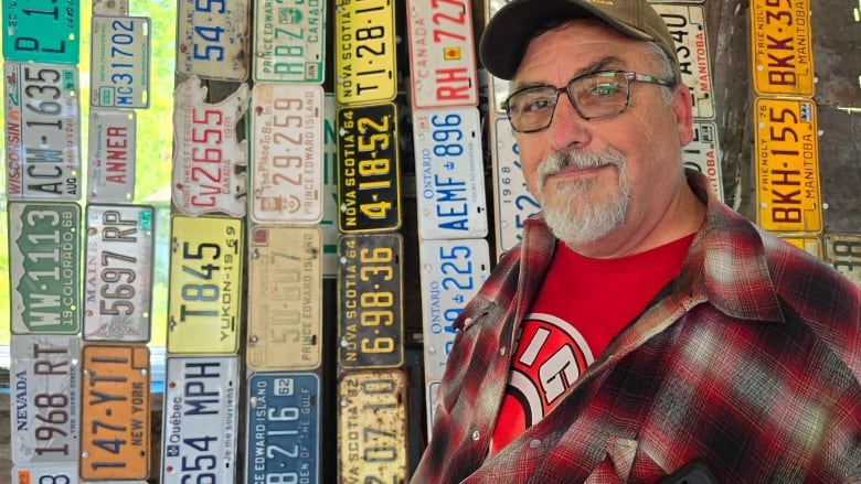 A man with a greying goatee, glasses and a baseball hate wears a red plaid jacket while standing in front of rows of different license plates from all over Canada.