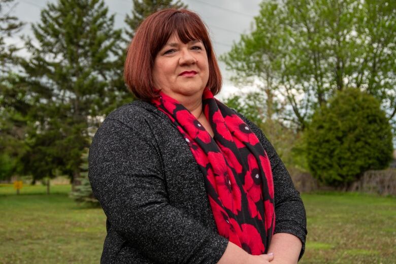 A person with medium-length red hair who is wearing a grey sweater and a black scarf with red poppies, stands in a green space.