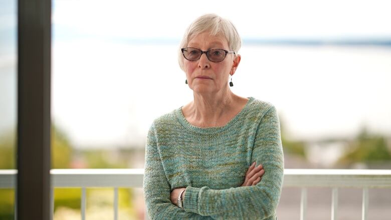 An older white woman wearing darkened glasses and a green and blue knit sweater stands on her balcony with her arms crossed. 