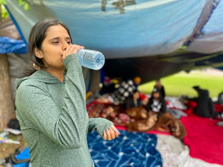 A woman with long, dark hair in a ponytail drinks from a water bottle in front of a tent encampment. 