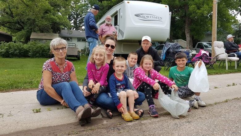 A woman sits on the edge of a sidewalk with a big group of adults and kids. 