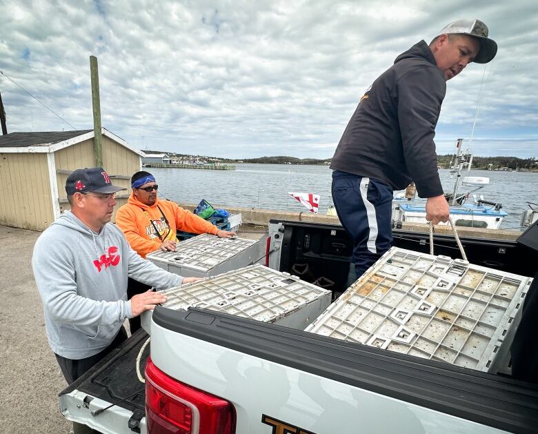 Two men push large plastic crates into the back of a pickup truck while a third man moves one by its rope handle, with a harbour in the background.