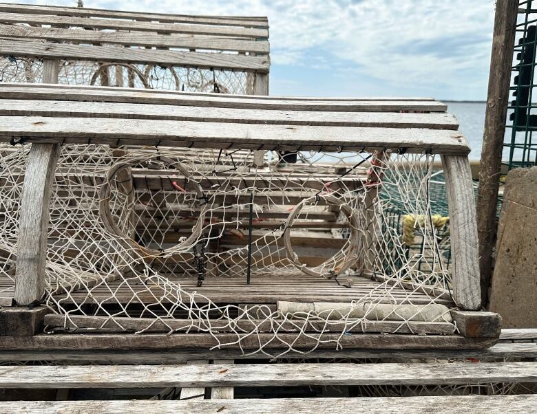 A wood-and-mesh lobster trap is seen in a stack on a wharf with the mesh cut open along the side.