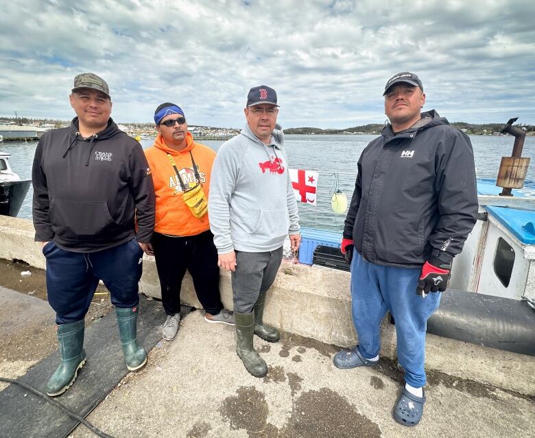 Four men in rubber boots and shoes wearing baseball caps and a bandana stand on a wharf with their boat and the harbour water in the background.