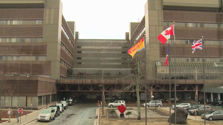 A large brown building with flags, parked cars and pedestrians out front.