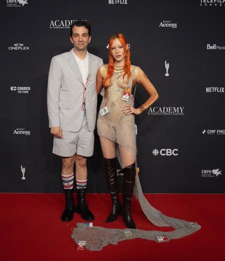 Jay Baruchel in a striped short suit and Rebecca-Jo Dunham-Baruchel in a crochet dress ayered over a bodysuit, embellished with cigarette packs.  