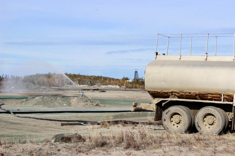 A water truck with a sprayer set up in in the background, wetting down a vast area of grey material.