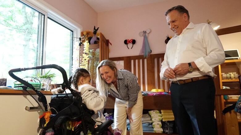 A smiling Marilyne Picard bending down toward a young girl in a type of wheelchair as Franois Legault looks on smiling. 