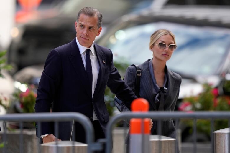 A cleanshaven middle-aged man in a suit and tie is shown walking outside with a woman wearing sunglasses and a blazer.