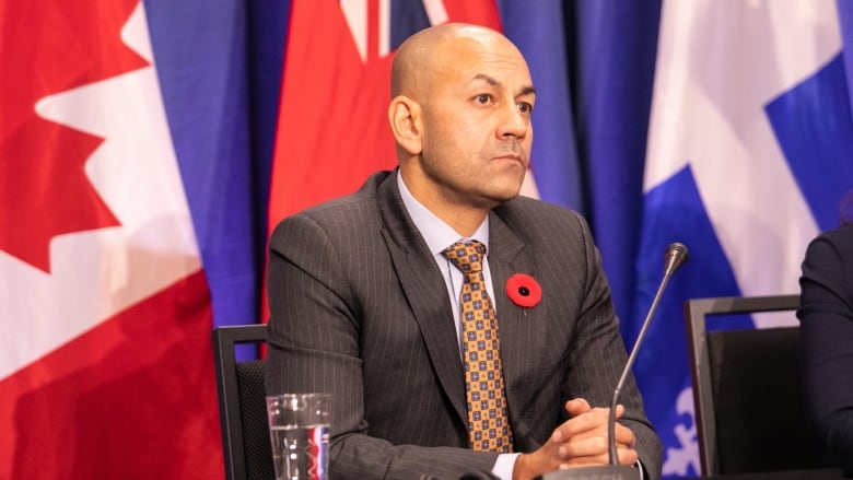 A man in a suit sits at a table before a microphone, with a backdrop of flags.