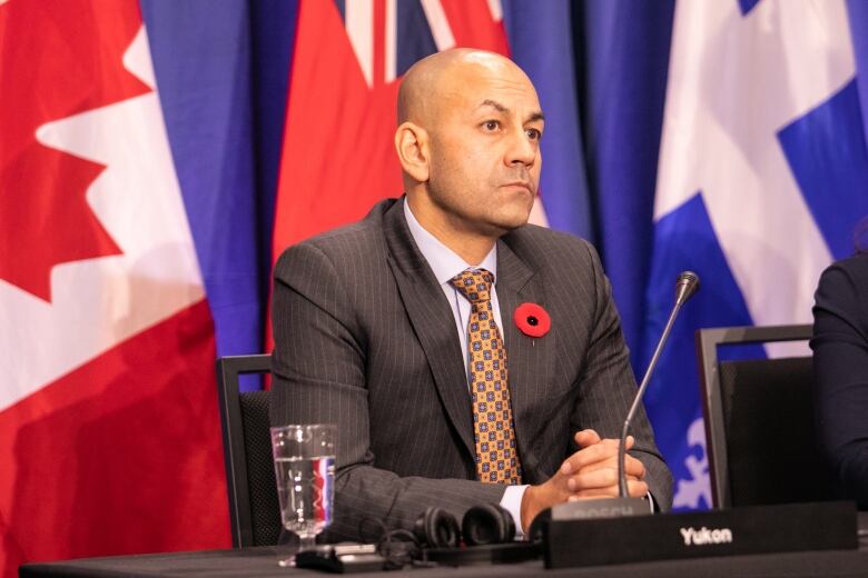 A man in a suit sits at a table before a microphone, with a backdrop of flags.