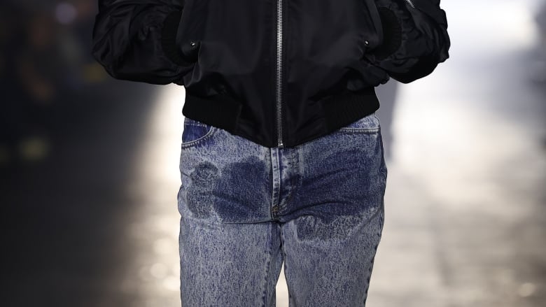 A man with oversized sunglasses, a black jacket and jeans with a stain in the front walks down a runway. IN the background, bright lights glow.