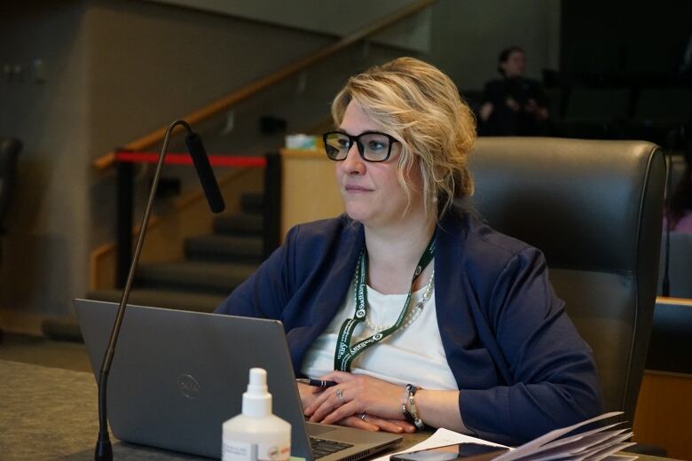 A blonde woman wearing black glasses sitting behind a laptop.