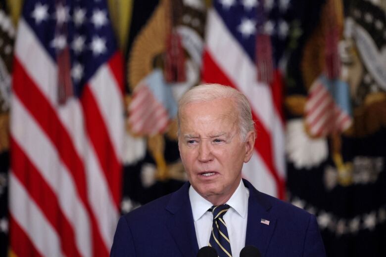 A man in a navy blue suit with a blue-and-yellow striped tie stands at a podium with American flags in the background.