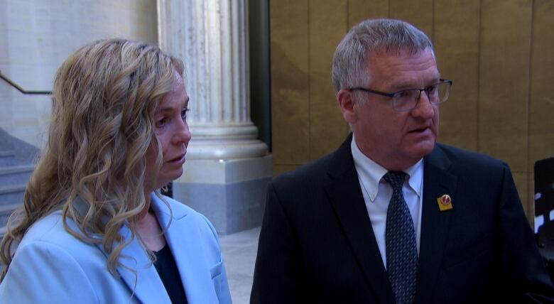 Woman with blue suit stands next to a man wearing a black suit. 