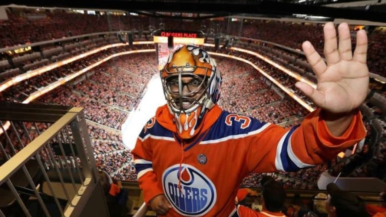 A man stands waving from the top seats at Rogers place, wearing a hockey goalie mask and an Oilers jersey. 