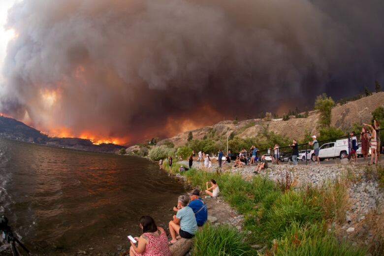 People stand and sit at the edge of body of water. A fire rages across the water and the sky is dark with smoke.