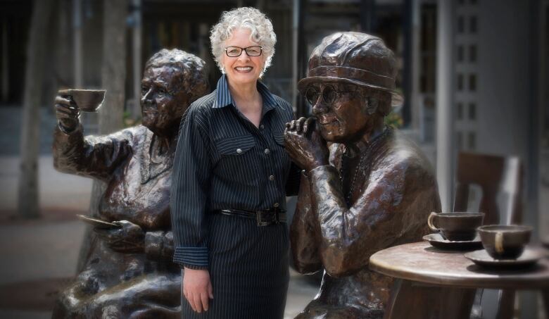 A white woman with short hair and black dress clothing stands between bronze sculptures on a sunny day.