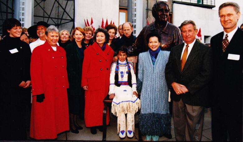 A group of people pose with a sculpture as a young girl sits in a bronze chair. 