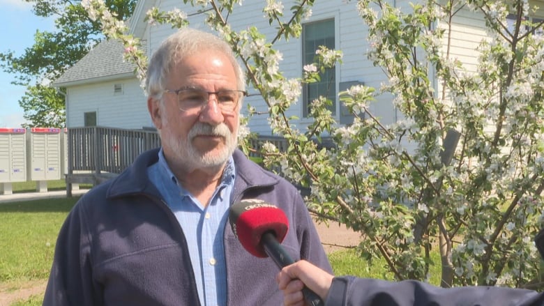 A man with white hair, a white beard and glasses stands for an interview with CBC News. There is a white building behind him with mailboxes and a tree blooming with flowers. 