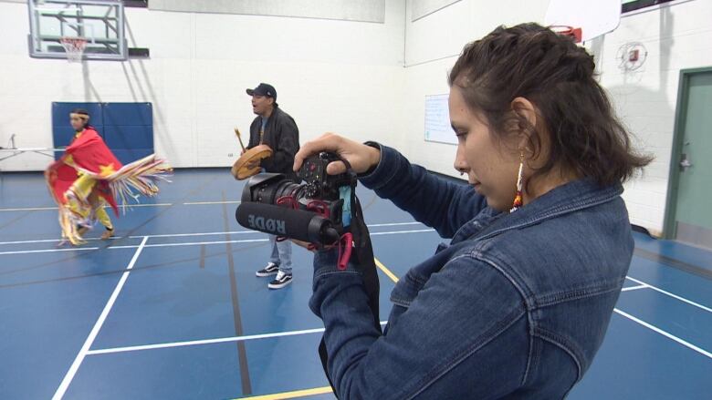 A woman with her hair in braids stands sideways to the camera, looking through a video camera of her own. A man plays a drum and a powwow dancer dances in the background.