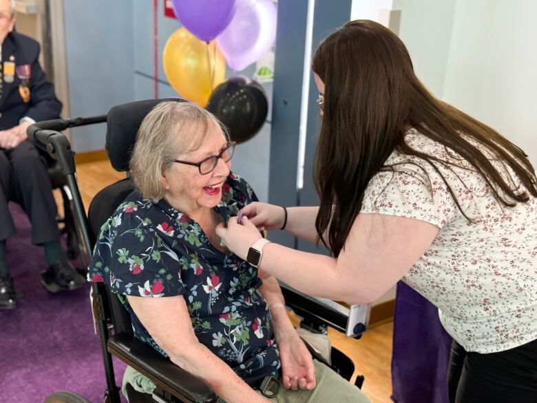 A 100-year-old woman in a wheelchair accepts a ribbon being pinned to her shirt by another woman. 