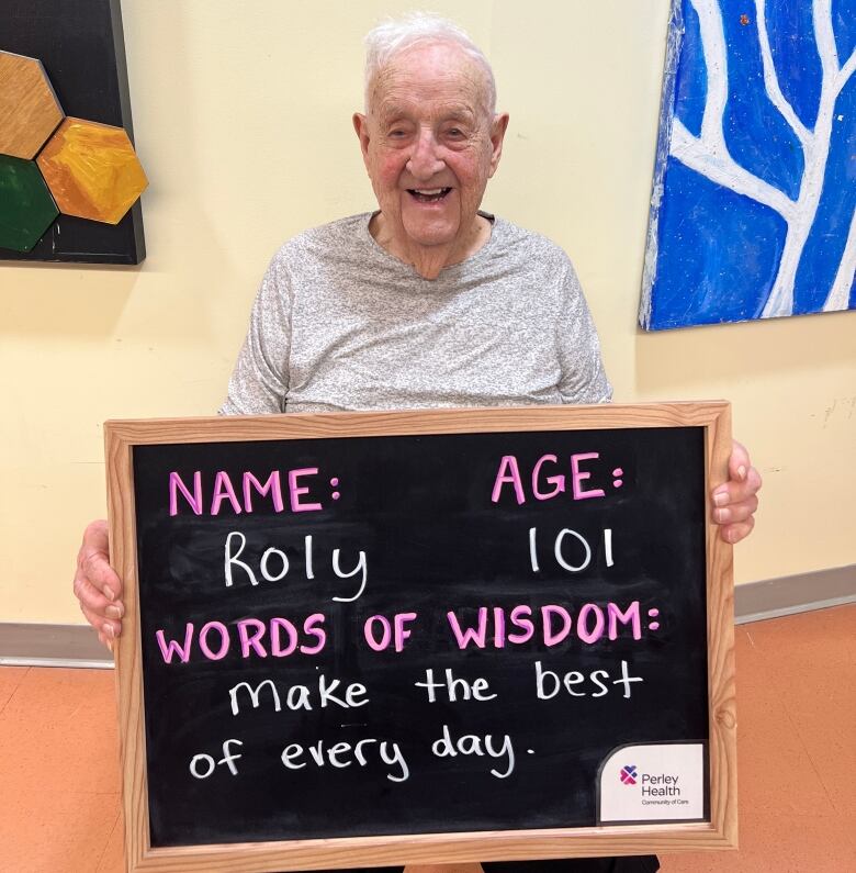 A man sits with a chalkboard that reads NAME: Roly AGE: 101 WORDS OF WISDOM: make the best of everyday. 