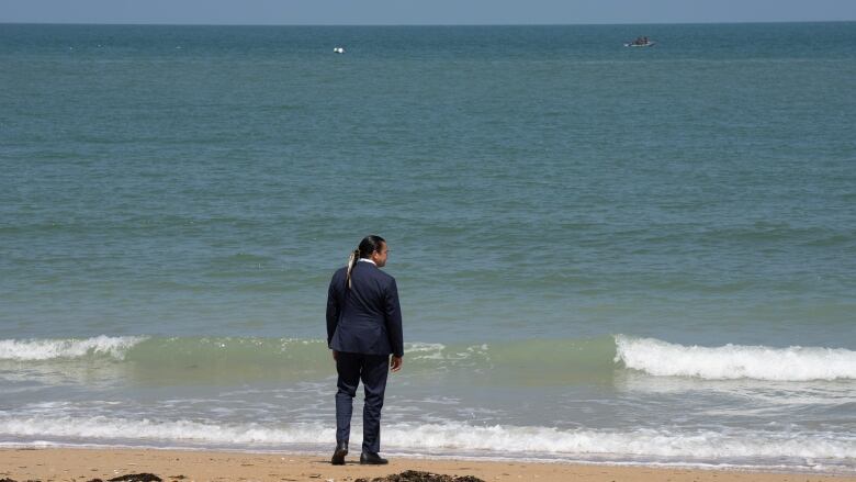 A man in a suit stands on a beach, looking out at the water.