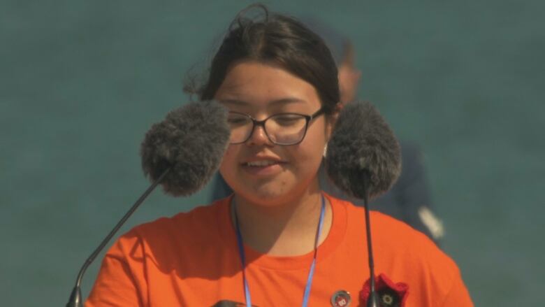 A girl in glasses and an orange T-shirt speaks into a microphone outside at a podium