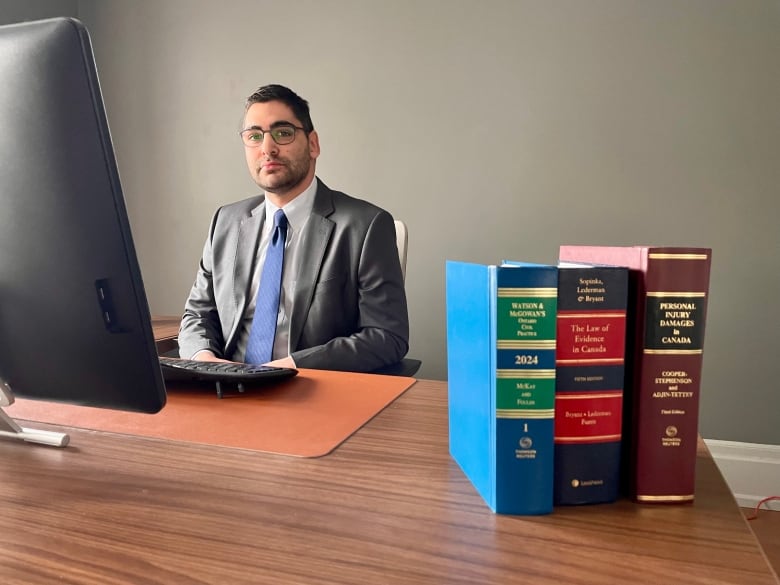 A dark-haired man wearing glasses, a grey suit and light blue tie sits at a desk in front of a computer monitor. Three thick law books sit on the desk in the foreground.