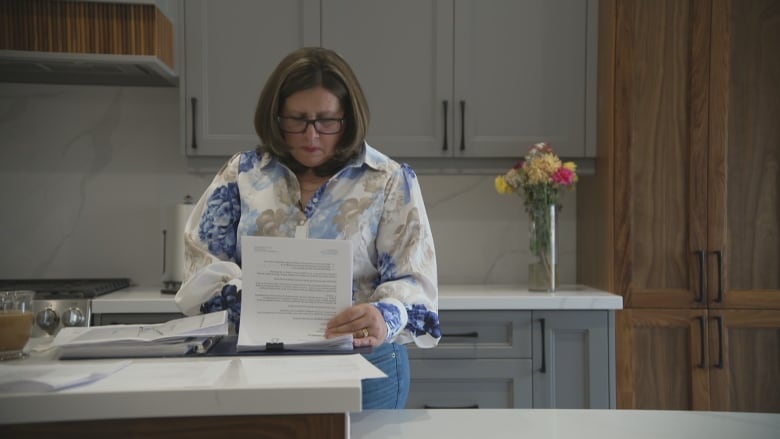 A woman with glasses and shoulder-length brown hair wearing blue jeans and a white print blouse with large blue flowers stands at a kitchen counter looking at a thick stack of papers in a file folder.