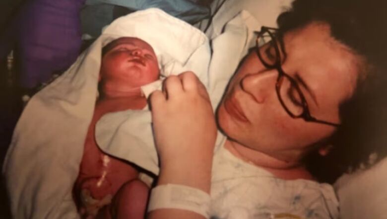 A dark-haired woman with glasses is reclined against a pillow holding a newborn baby.