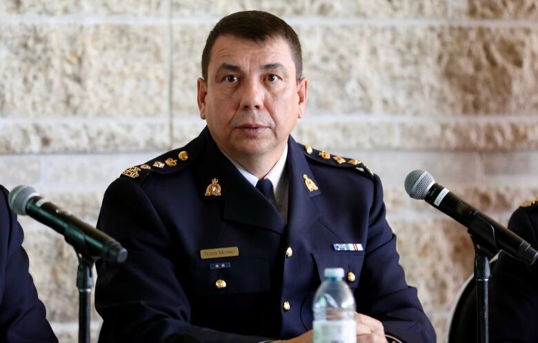 A man in a blue dress uniform sits at a table where there are two microphones. 