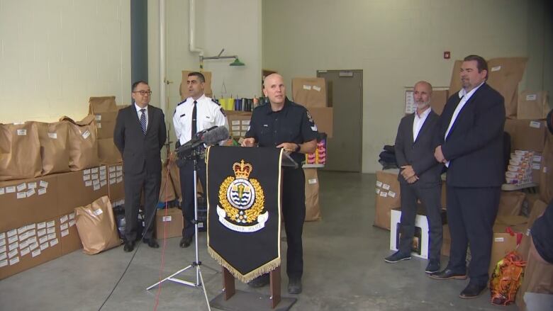 A man in a VPD uniform flanked by police officials and surrounded by boxes and bags speaks from a podium emblazoned with a Vancouver police crest.