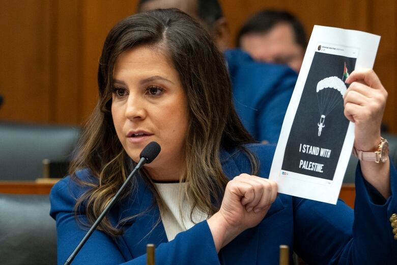 A dark-haired woman seated in a chamber wearing a blazer holds up a piece of paper that has a drawing and the phrase of 'I Stand With Palestine.'