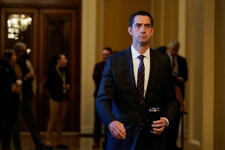 A cleanshaven dark haired man in a suit and tie holds a beverage container while standing in a hallway.