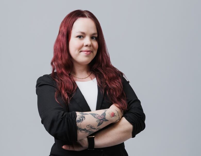 A woman with long red hair and a black blazer crosses her arms in a pose while smiling. 